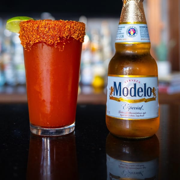 A Modelo beer bottle next to a michelada with a chili-lime rim and a lime wedge.